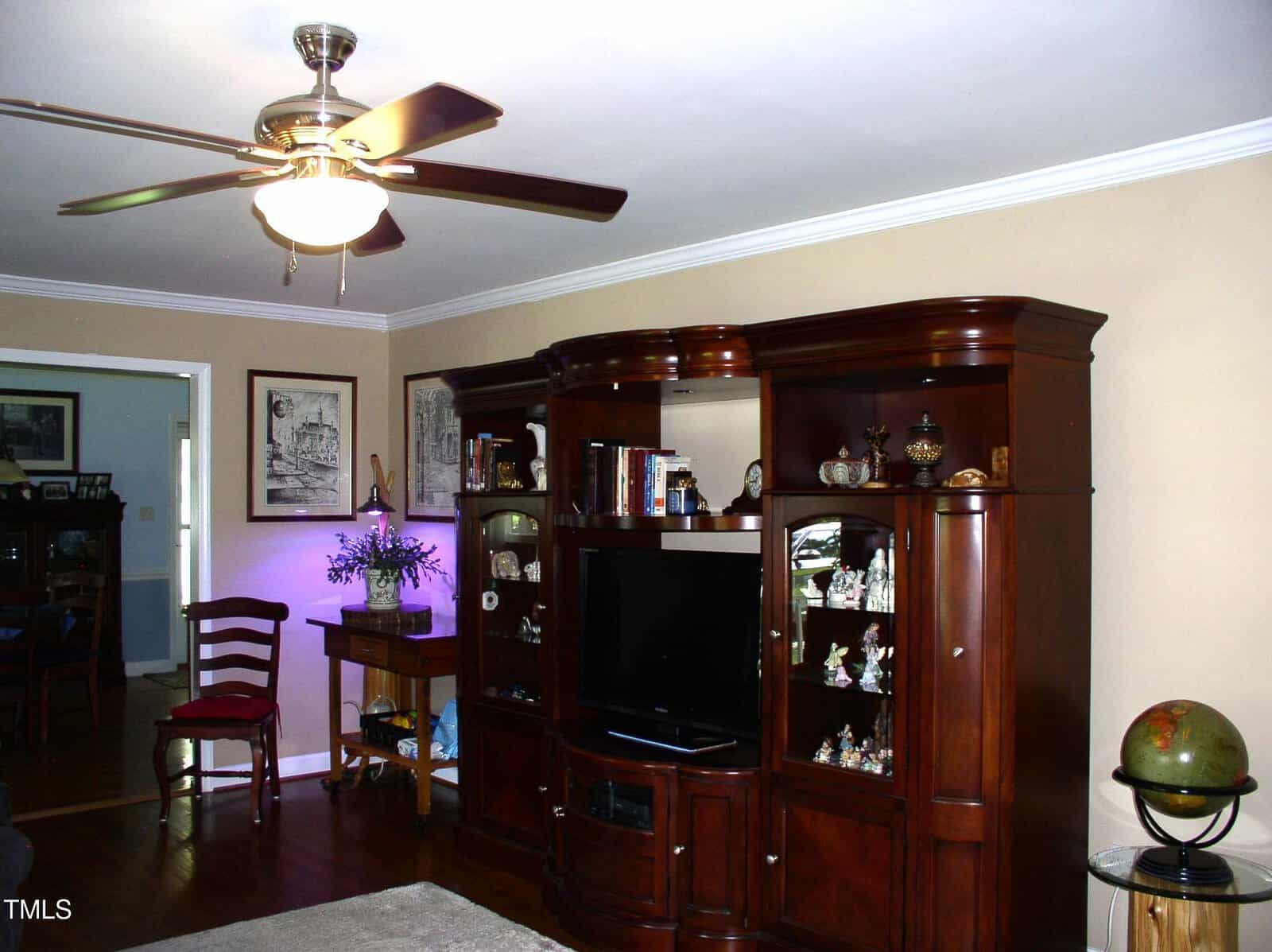 A living room with wooden floors, a ceiling fan with a light, a large dark wood entertainment center holding a TV and decor, two red chairs, a small table with a lamp, and a globe on the right.