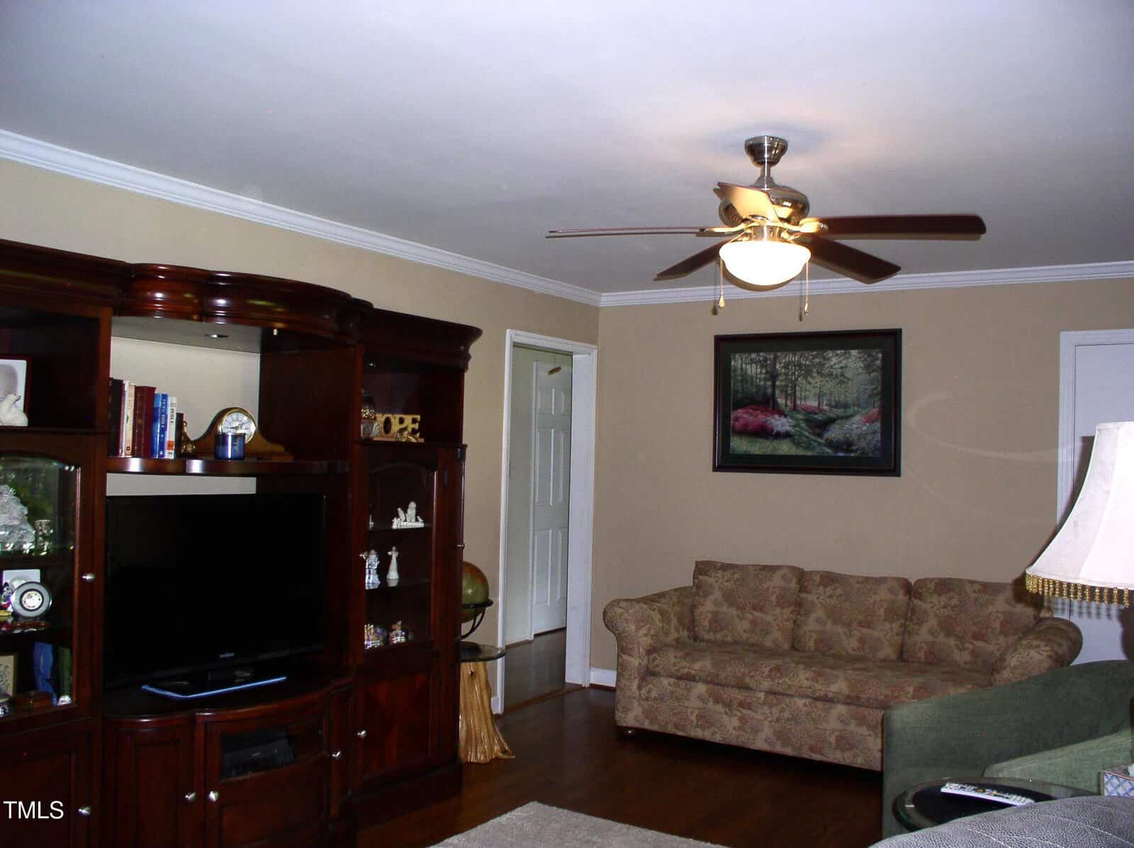 A living room with beige walls, a floral-patterned sofa, a green armchair, a dark wood entertainment center with a TV and shelves, a ceiling fan with a light, a floor lamp, and a landscape painting on the wall.