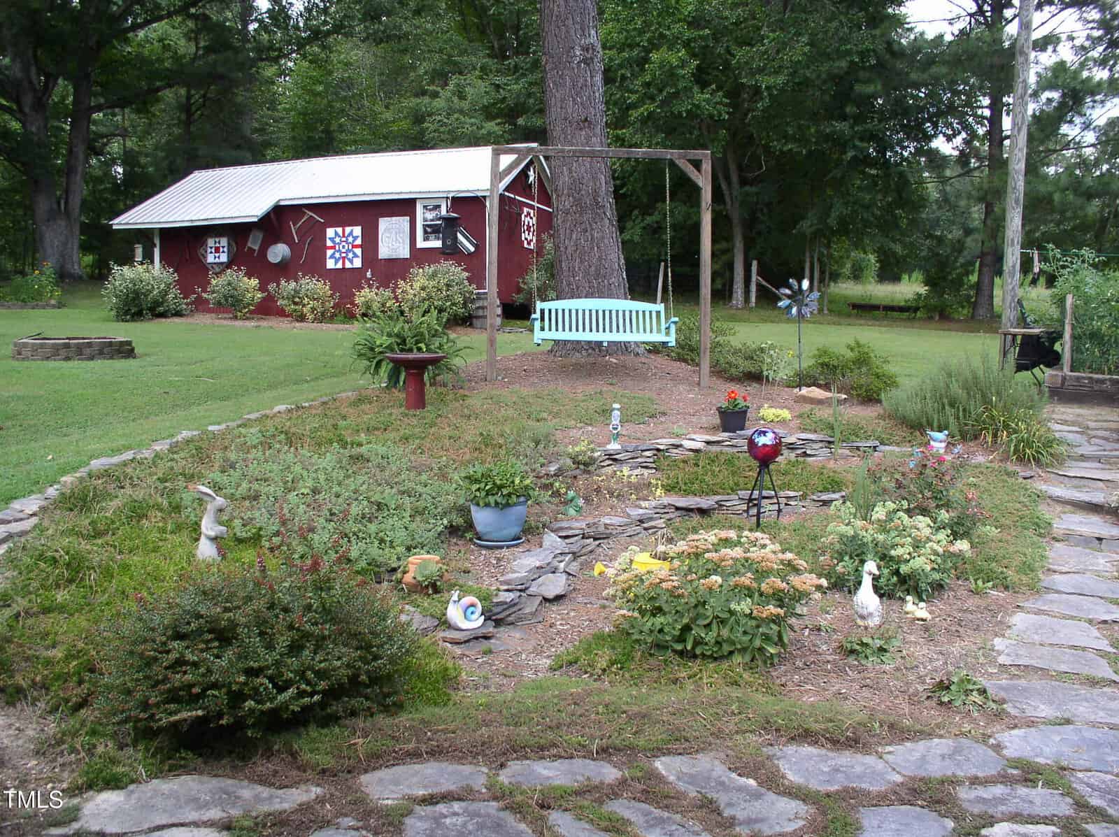 A landscaped garden with a variety of plants, lawn ornaments, and a small pond; a light blue bench sits under a wooden arbor, and a red shed with white trim is in the background surrounded by trees.
