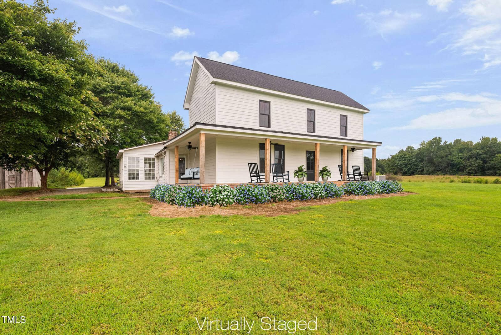 A two-story white house with black roof and shutters at 8365 Aaron Creek Church Road, Oxford. Features a wrap-around porch with rocking chairs, surrounded by grass and landscaping, trees, and a blue sky. Text at the bottom reads "Virtually Staged.