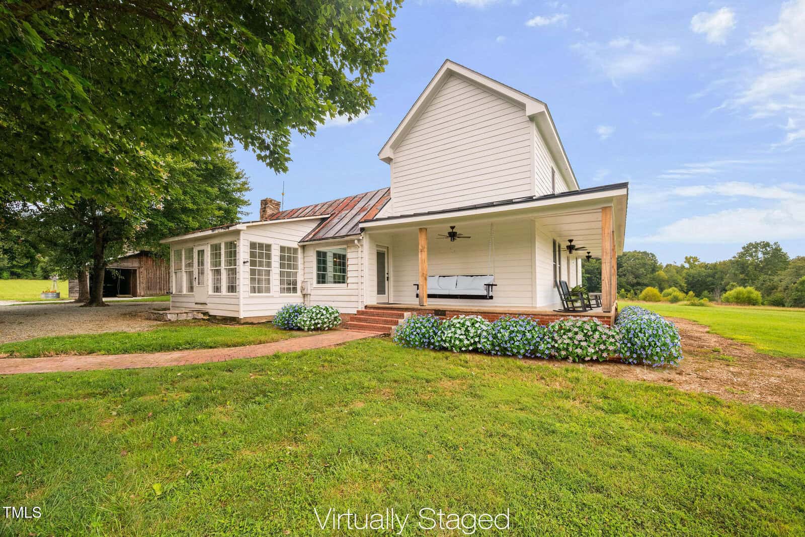 A white two-story farmhouse with a front porch, ceiling fans, and hydrangea bushes, set on a grassy lawn with trees and an open field in the background. The image is labeled "Virtually Staged.