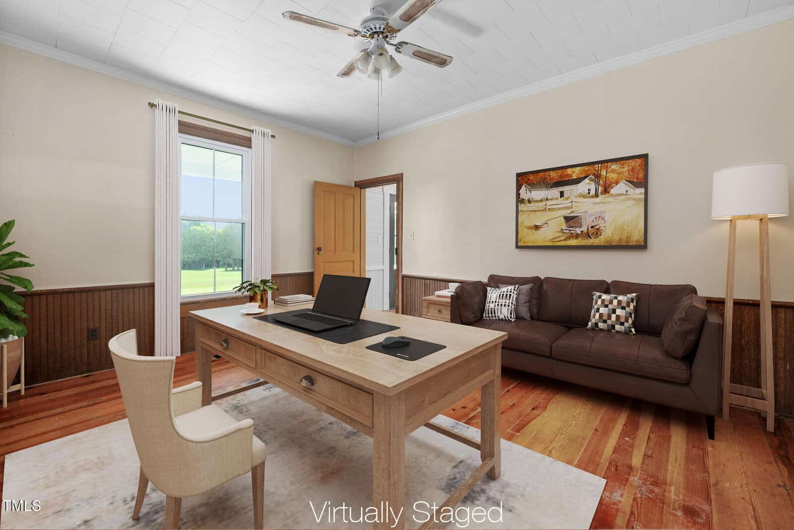 A home office features a wooden desk with a laptop, chair, and mouse on a light rug. A brown sofa, floor lamp, and artwork are against the wall. Sunlight enters through a window with white curtains. "Virtually Staged" text at bottom.