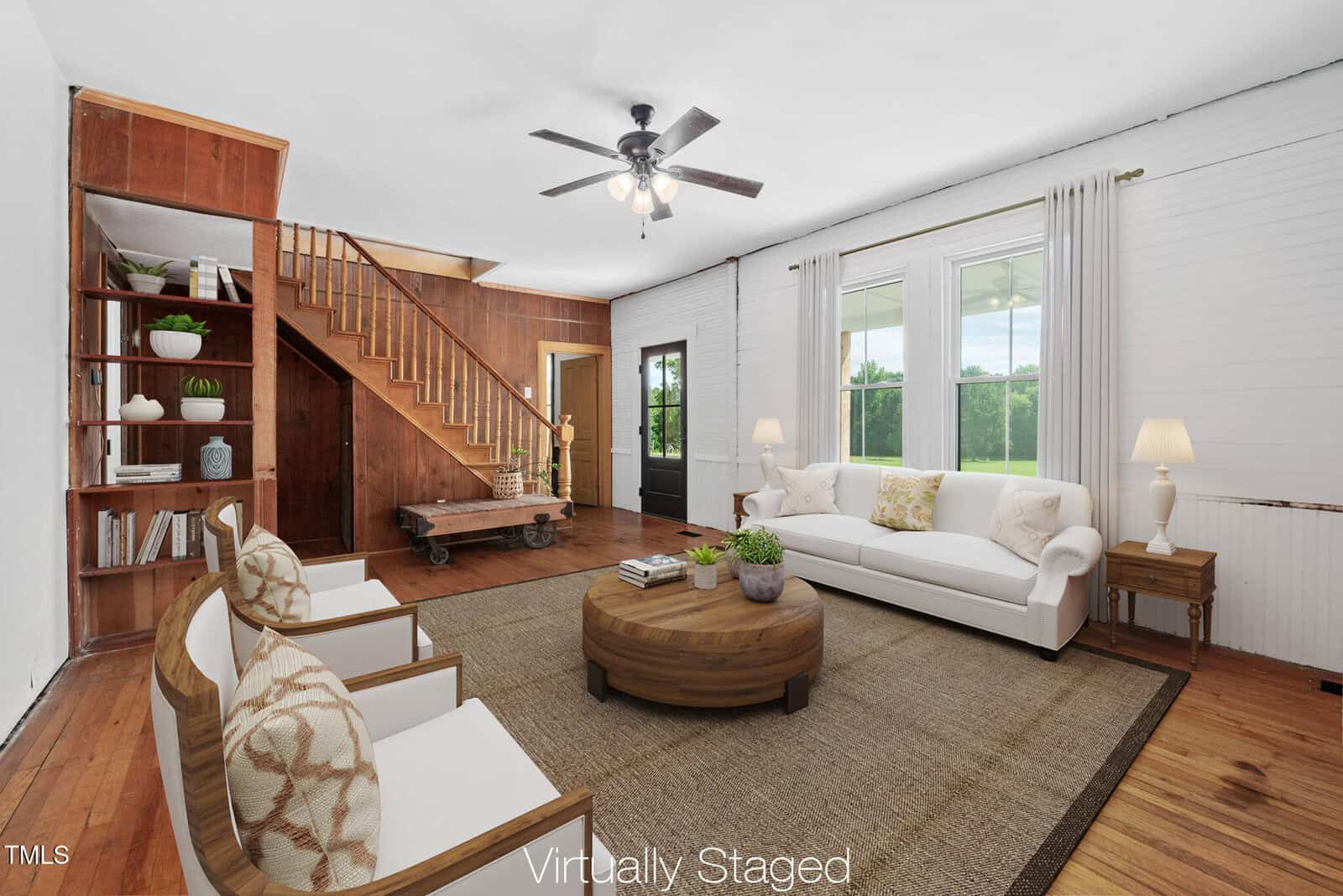A virtually staged living room with wood floors, a white sofa, two white armchairs, a round coffee table, and built-in shelves. Wooden stairs lead to an upper level, and large windows let in natural light.
