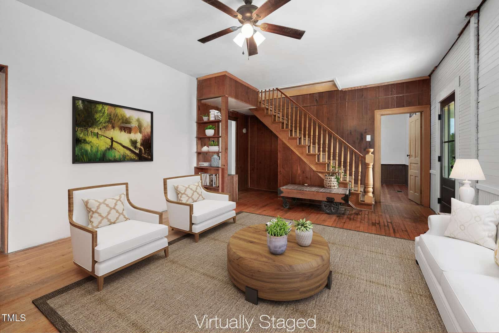 A living room with a wooden staircase, white walls, wood-paneled accent wall, and ceiling fan. Virtually staged with white armchairs, a white sofa, a round wooden coffee table, and wall art. A rug covers the floor.