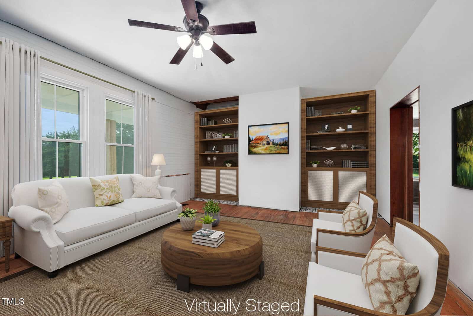 A living room with white walls, a ceiling fan, two windows, a white sofa, two armchairs, a round wooden coffee table, built-in bookshelves, a painting, and the text "Virtually Staged" at the bottom.