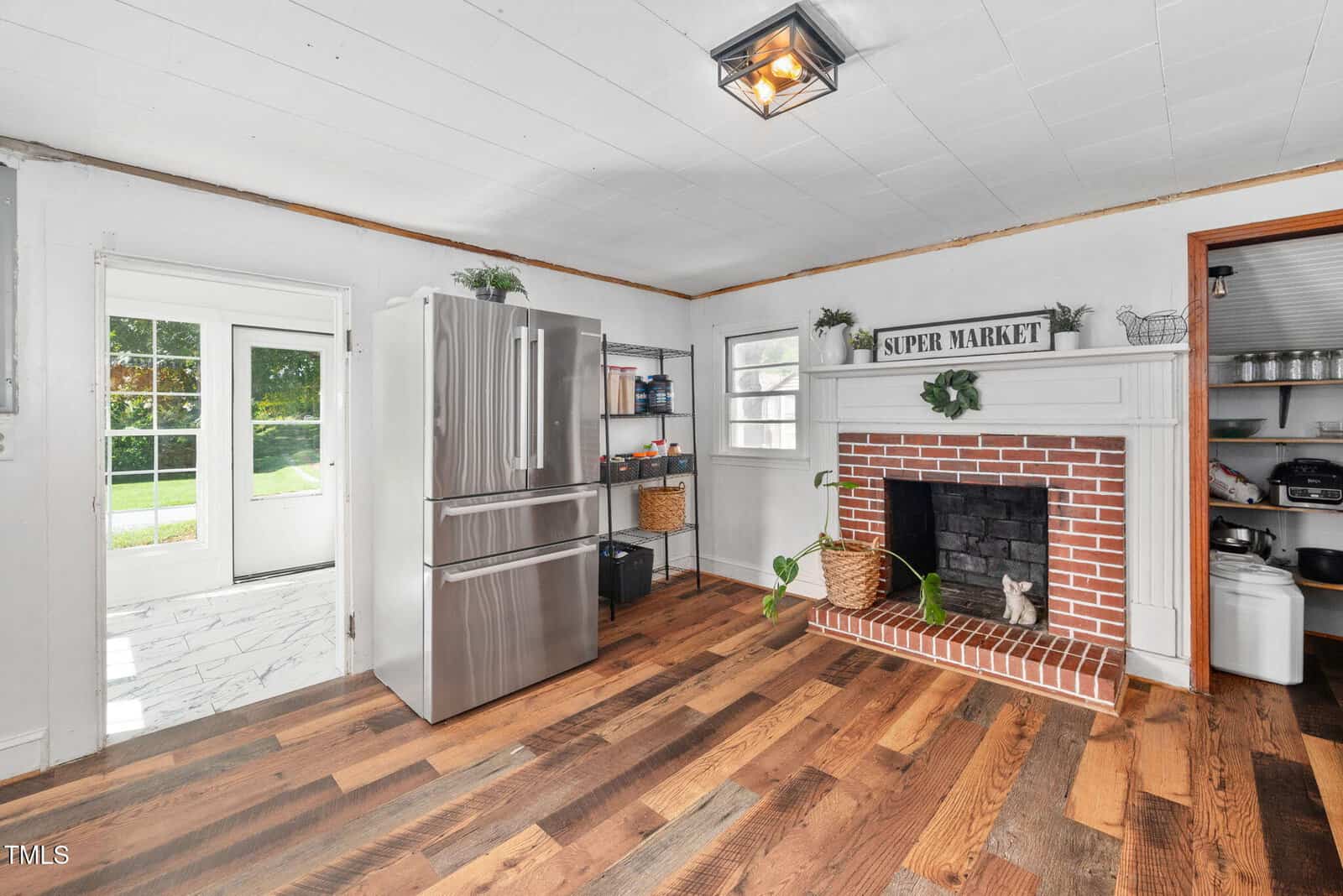 A kitchen with a stainless steel refrigerator, wood flooring, a white brick fireplace with a "Super Market" sign on the mantel, and a pantry visible through an open door. Natural light enters through glass doors.