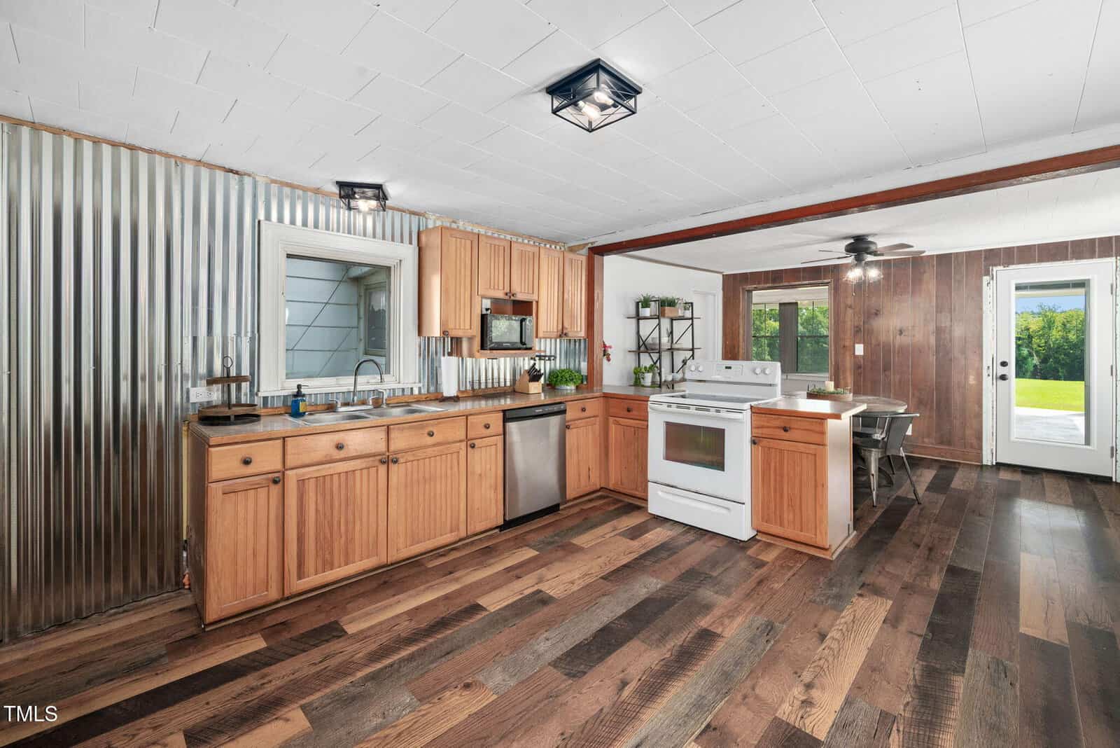 Rustic kitchen with wood cabinets, corrugated metal backsplash, stainless steel appliances, white stove, wood-paneled wall, large window, glass door, and dark wood flooring. Ceiling lights give a modern touch.