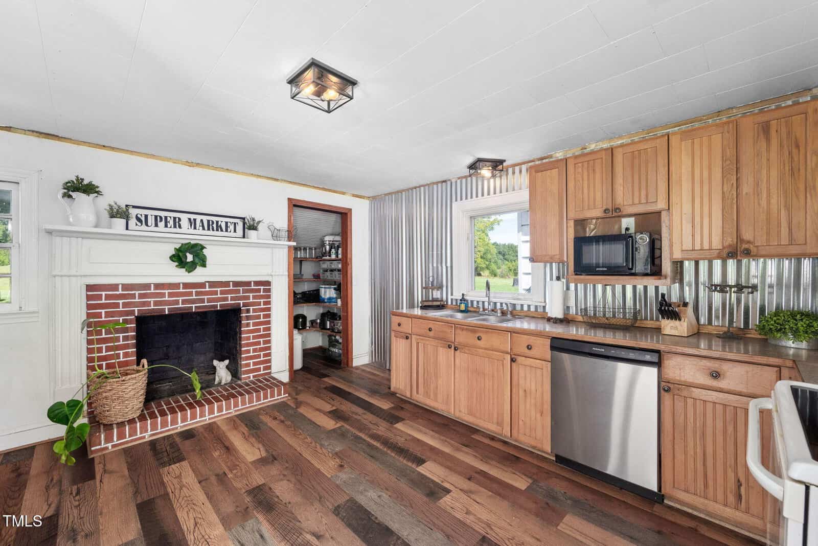 A kitchen with wood cabinets, stainless steel appliances, corrugated metal backsplash, and dark wood floors. There is a red brick fireplace with a "Super Market" sign above and a pantry visible through an open doorway.