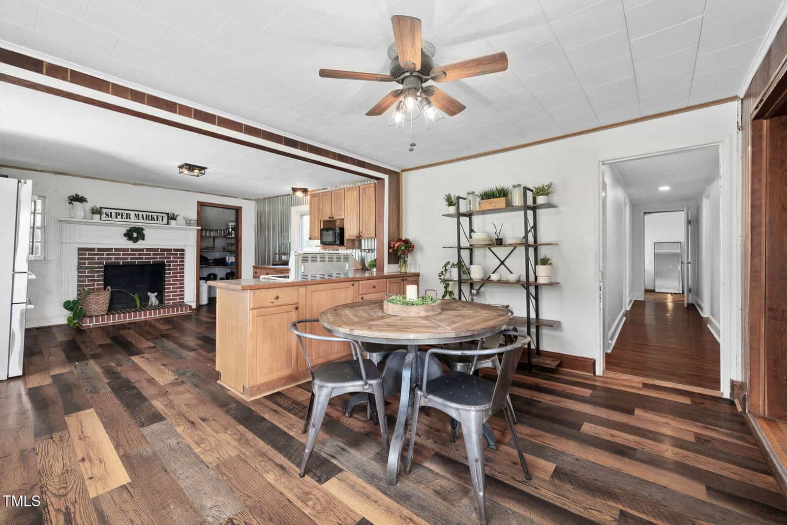 Open kitchen and dining area with wood floors, round table and metal chairs. Fireplace with “Super Mart” sign, ceiling fan, open shelving, hallway leading to other rooms, and natural light.