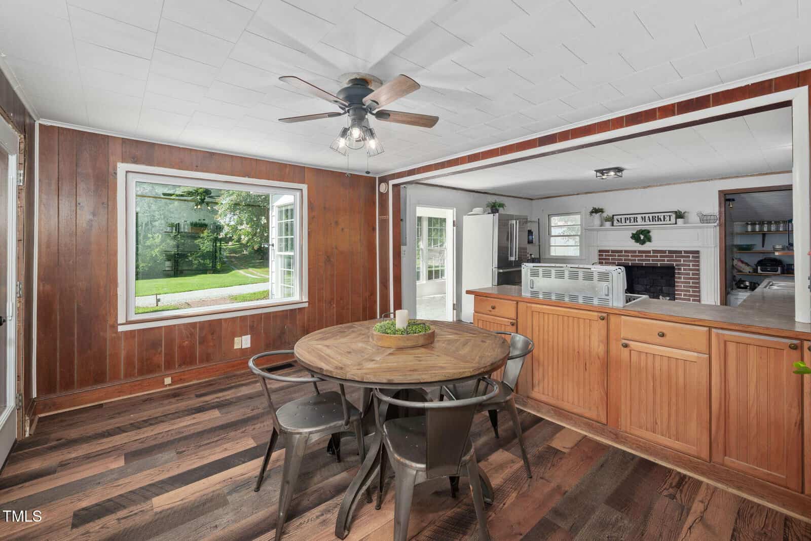 Dining area with a round wooden table, four metal chairs, wood-paneled walls, and large window. An open counter leads to a kitchen with white cabinets and a brick accent. Ceiling fan and natural light are visible.
