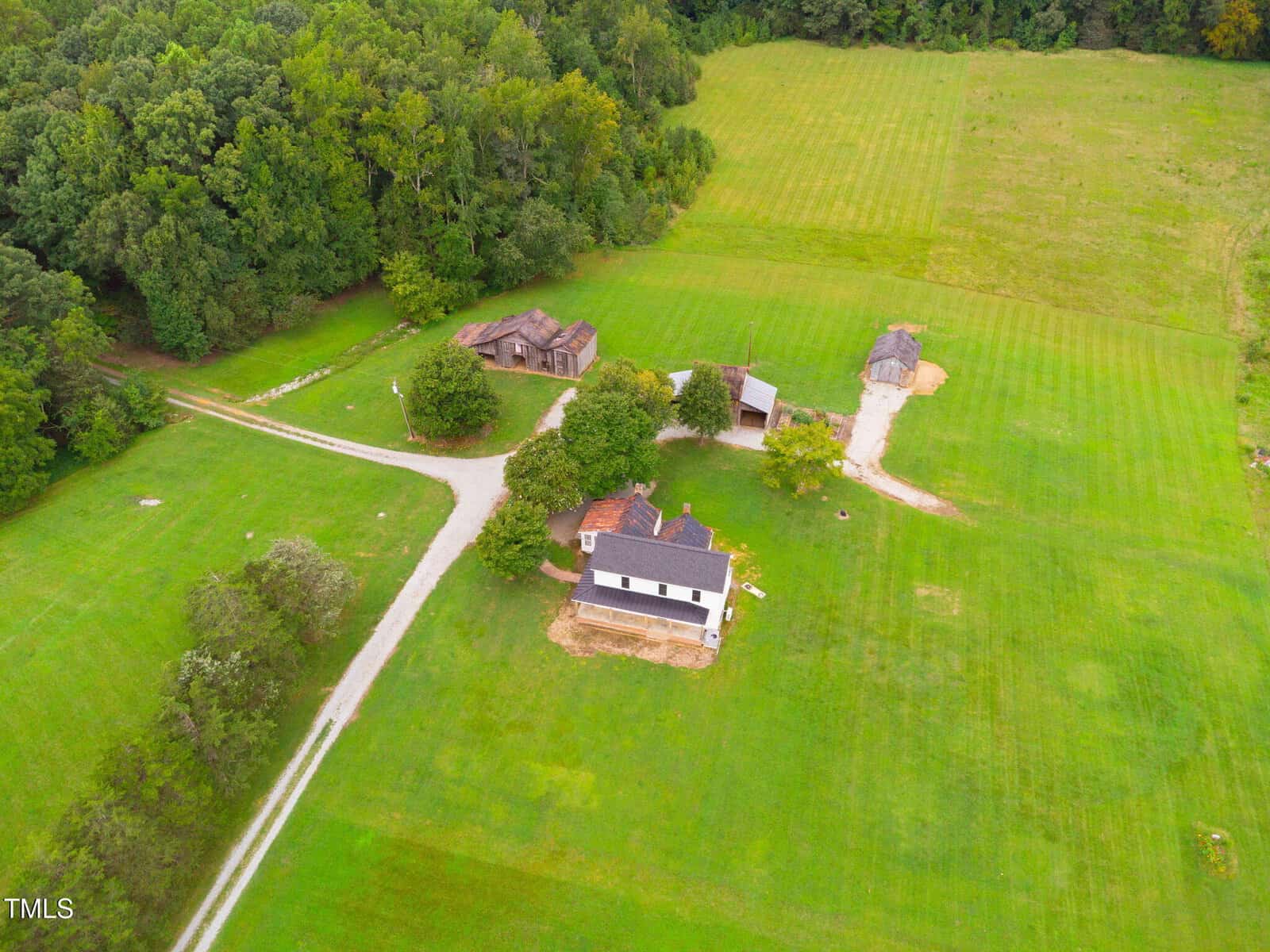 Aerial view of a rural property with a white house, several outbuildings, gravel driveways, and large grassy fields surrounded by dense trees.