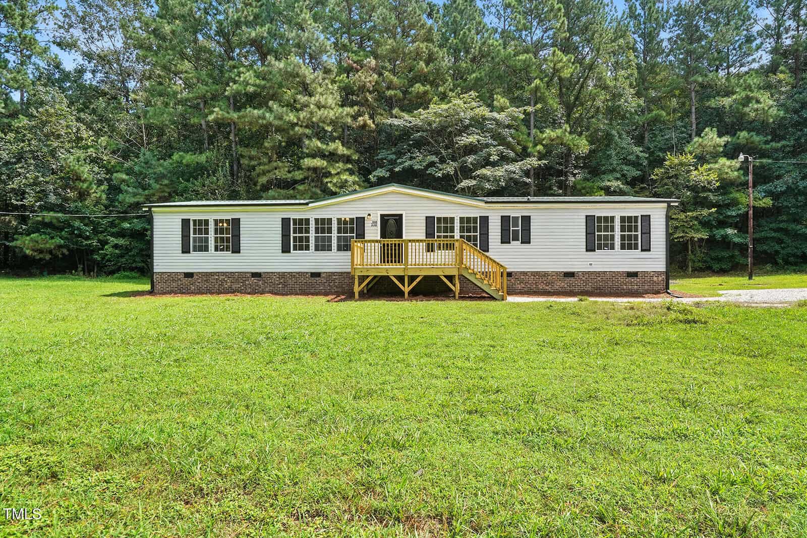 A single-story manufactured home at 330 Sycamore Lane in Henderson features beige siding, black shutters, a small yellow front porch with steps, a spacious grassy yard, and dense trees in the background.