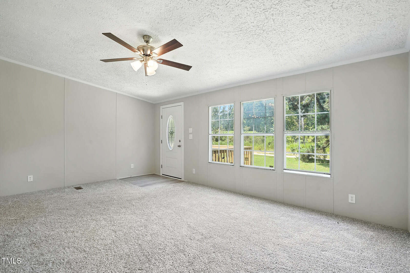 A carpeted living room with light gray walls, a white ceiling fan with lights, three large windows showing green trees outside, and a white front door with an oval window.