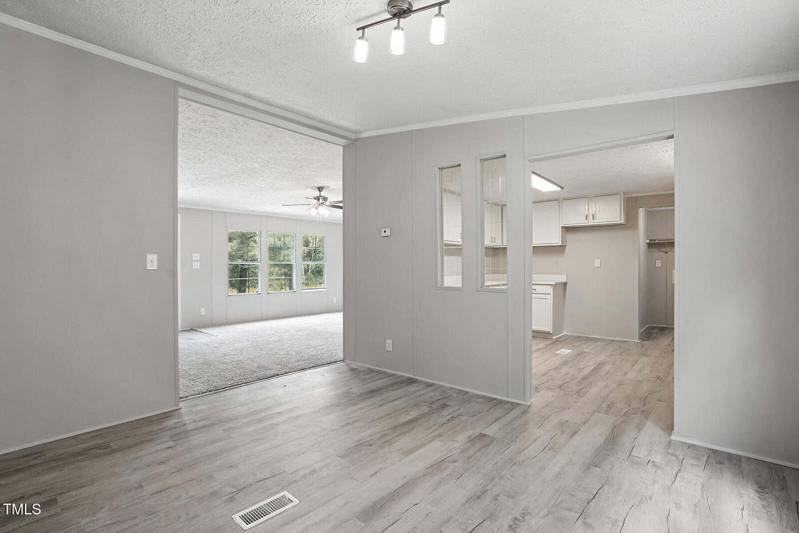A view of a modern, unfurnished open-plan home interior with light gray walls, wood-look flooring, and white trim. The space includes a living area, dining area, and a kitchen with white cabinets. Large windows provide natural light.