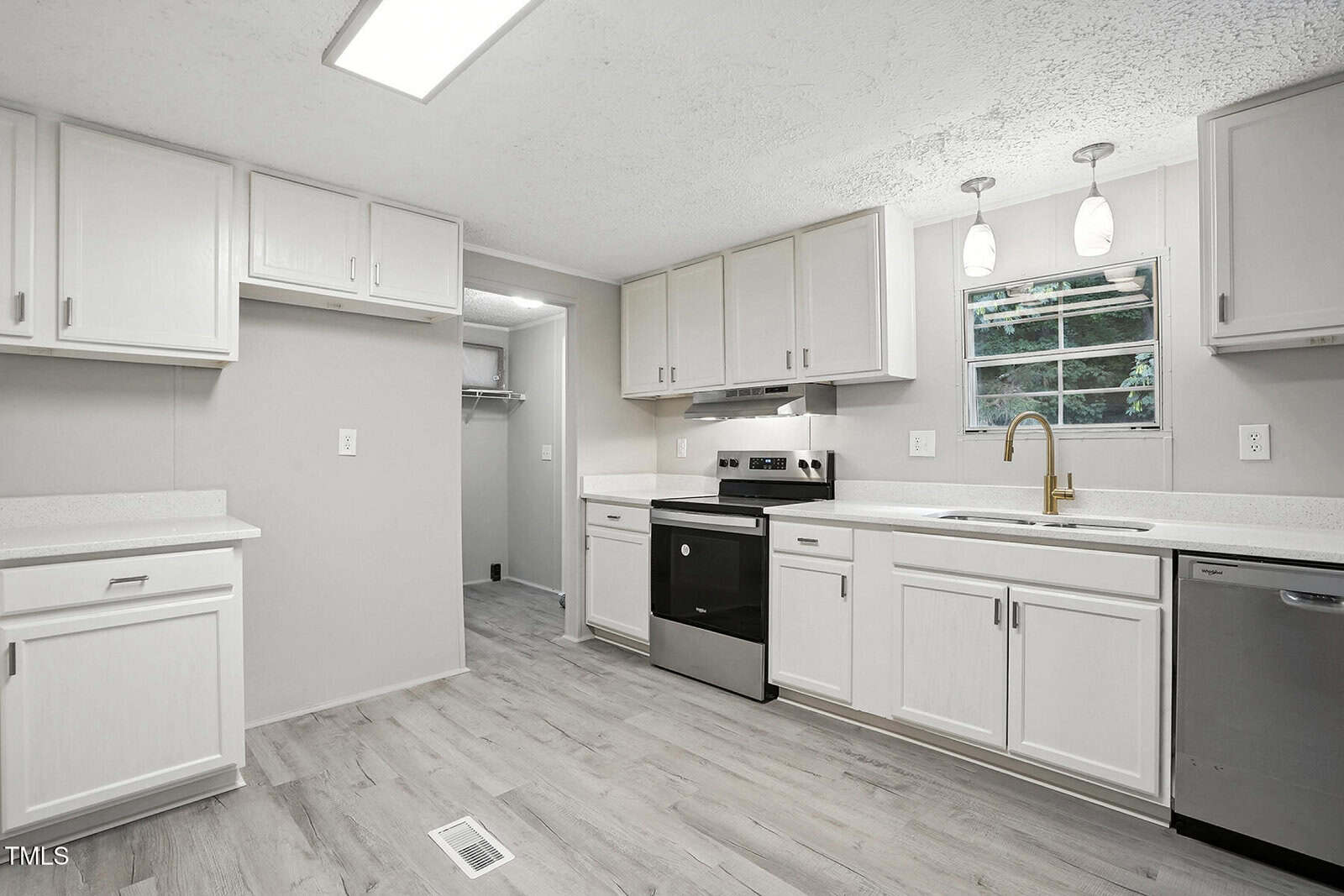 A modern kitchen with white cabinets, light gray walls, light wood flooring, stainless steel appliances, a window above the sink, and recessed lighting. An open doorway leads to a pantry or closet.