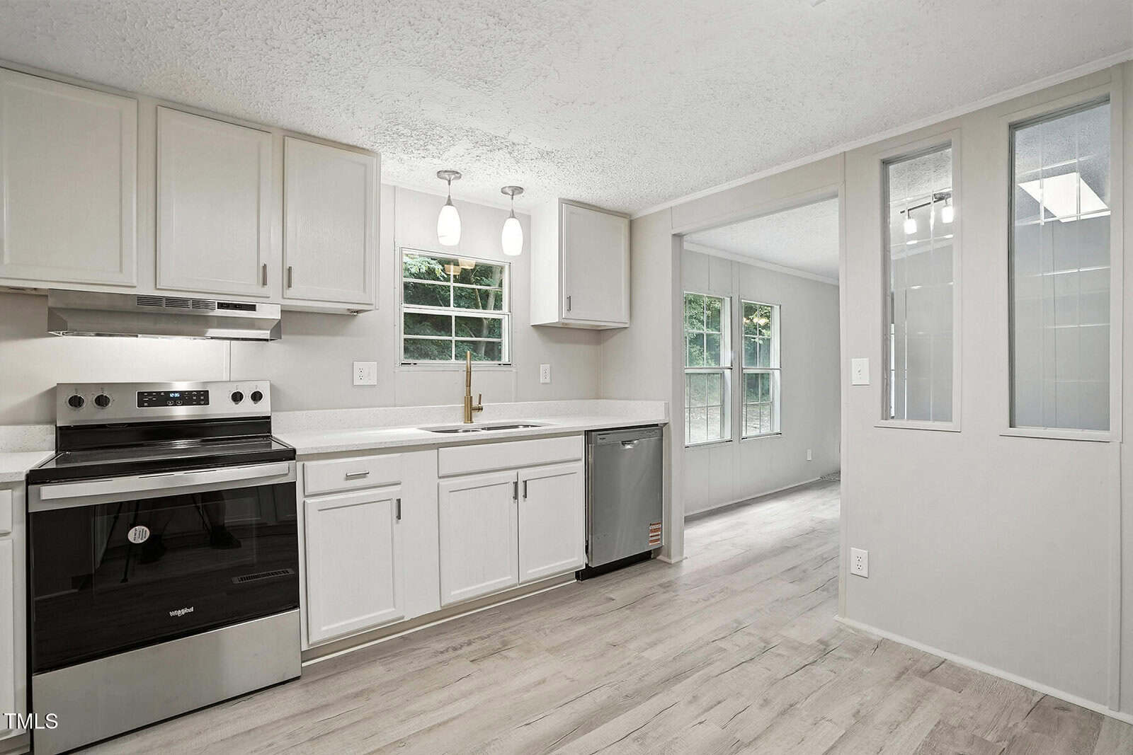 A modern kitchen with white cabinets, stainless steel oven and dishwasher, light countertops, two pendant lights, and windows overlooking greenery. Light wood flooring extends into an adjacent room.
