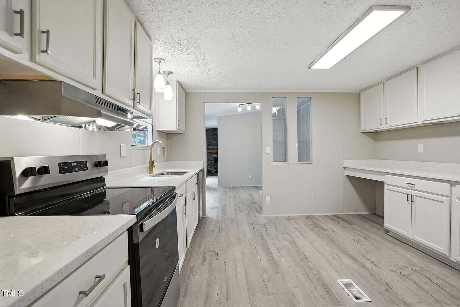 Modern kitchen with white cabinets, stainless steel appliances, light wood flooring, and quartz countertops. A window reveals an adjacent room with gray walls and a glimpse of another space beyond. Bright, clean, and spacious.