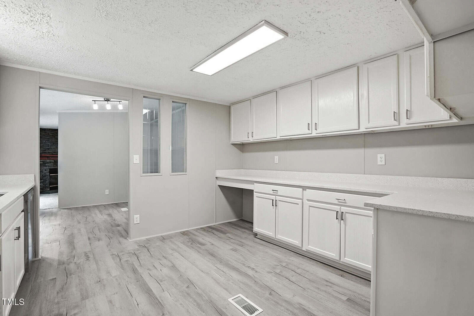 A spacious, modern kitchen with white cabinets, light gray walls, wood-look flooring, and bright overhead lighting. A large countertop runs beneath the cabinets. An open doorway leads to another room with a fireplace.