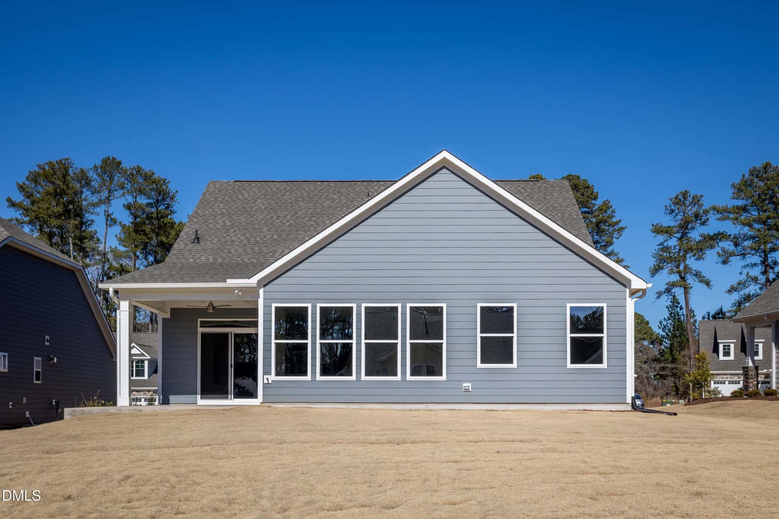 A modern, single-story house with blue siding, white trim, multiple windows, and a covered back patio, set on a dry grass lawn with trees and neighboring homes in the background under a clear blue sky.