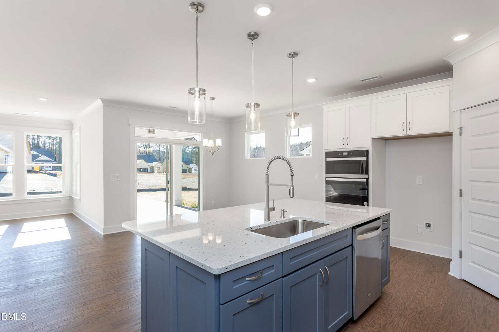 Modern kitchen with a blue island, white quartz countertop, stainless steel sink, and built-in oven. Pendant lights hang above the island, and large windows let in natural light, illuminating the wood floors.