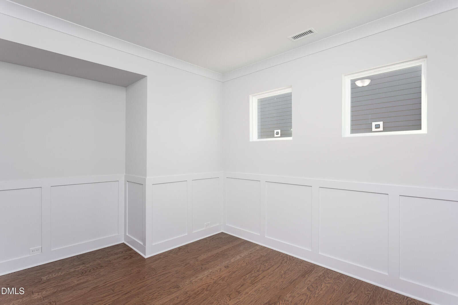 A bright, empty room with white paneled walls, two small windows, wood flooring, and a recessed wall area; ceiling vent and electrical outlets are visible.