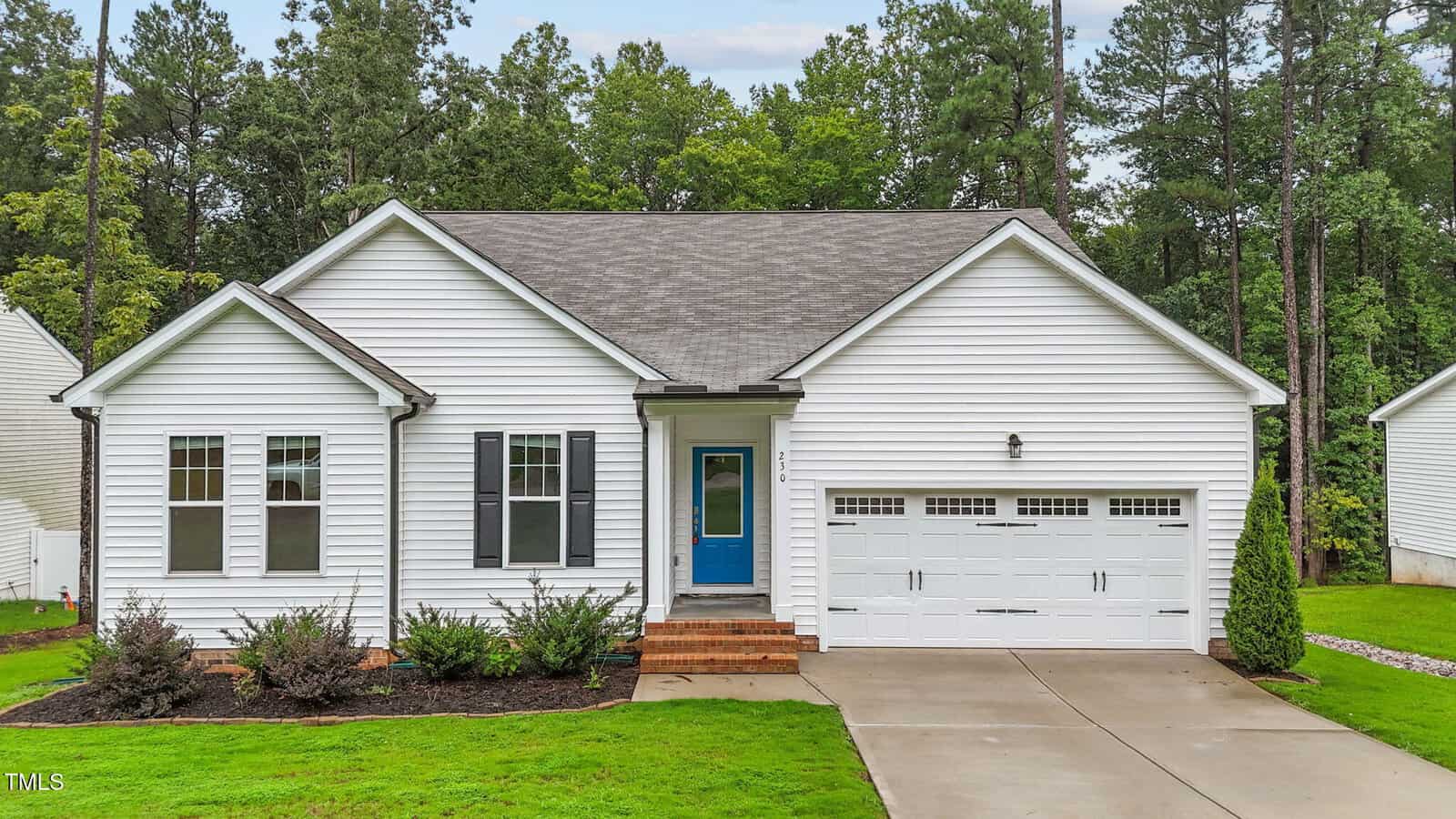 Single-story white house with a gray shingle roof, a blue front door, and a double garage located at 230 Sacred Fire Road in Louisburg. Brick steps lead to the entrance, with shrubs along the front and a concrete driveway. Tall trees and grass surround the property.