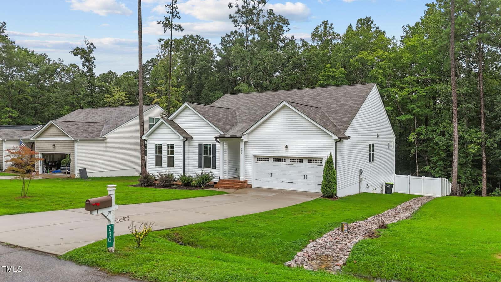 Single-story white house with a two-car garage, gabled roof, and front porch. Driveway and mailbox in front, with a grassy lawn and a rock-lined drainage ditch. Trees and neighboring houses in the background.