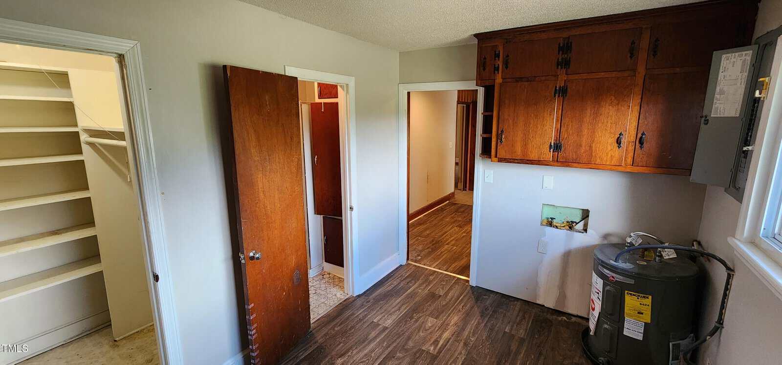 A small utility room with wood cabinets, a water heater, a brown door, and doorway leading to a pantry with shelves. The floor is wood-patterned, and the walls are light-colored. A window is visible on the right.