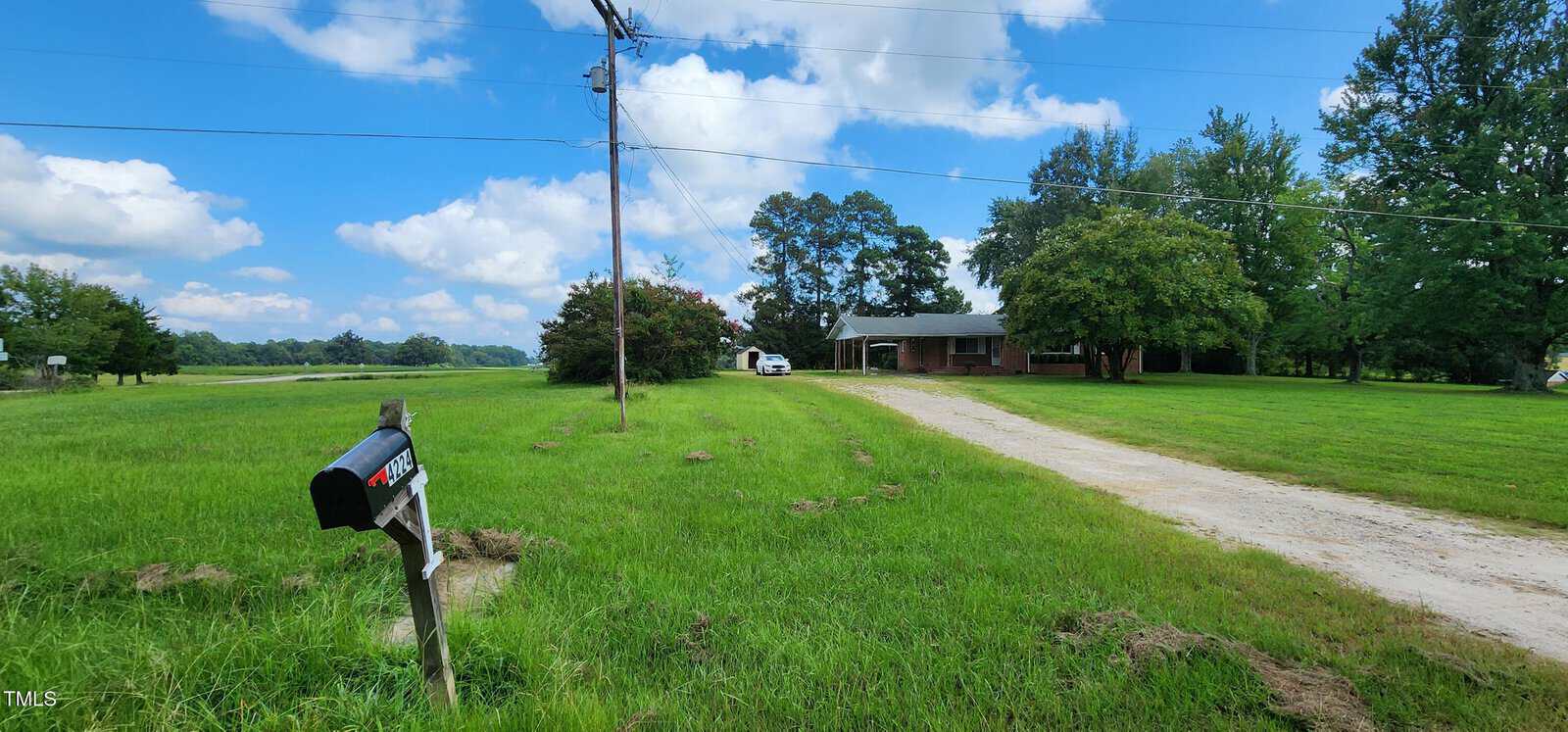 A mailbox labeled “4172” stands beside a gravel driveway leading to a single-story brick house surrounded by green grass, trees, and a partly cloudy sky in the quiet town of Louisburg. A white van is parked near the house, which is located not far from 4224 Us 401.