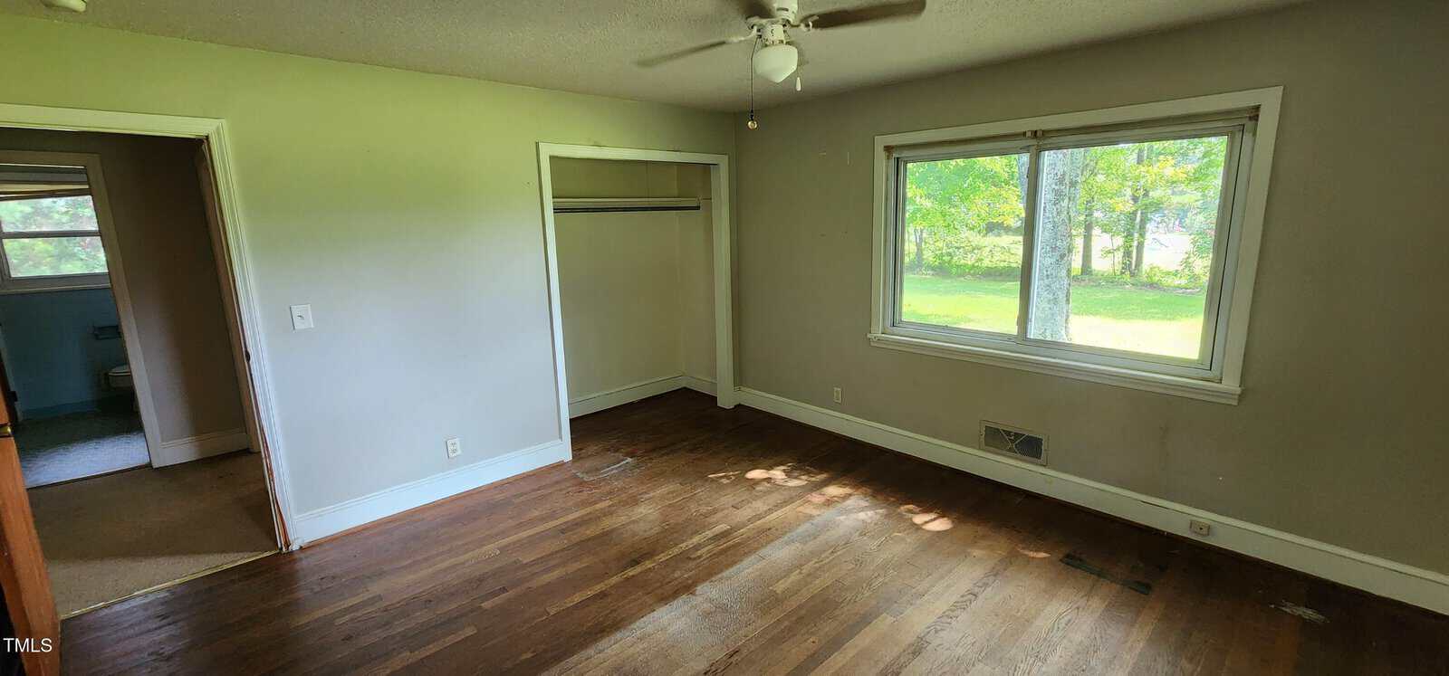 Empty room with hardwood floors, a ceiling fan, a window showing green trees outside, an open closet, light-colored walls, and a door leading to a bathroom. Sunlight is coming in, and the room appears clean but worn.