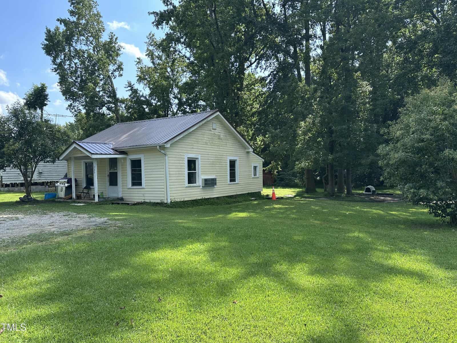 A small, single-story light yellow house with a metal roof sits on a grassy lawn, surrounded by tall trees and partial shade on a sunny day. A gravel driveway is visible to the left of the house.