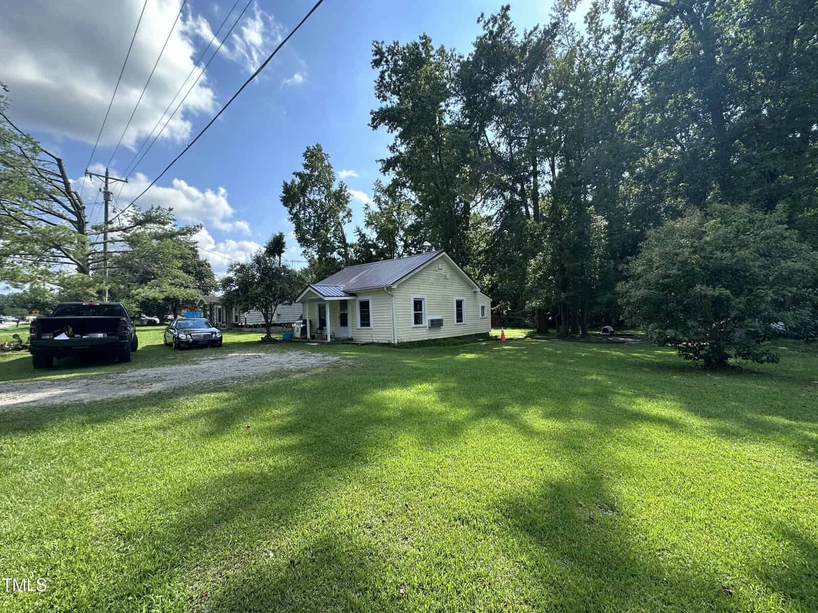 A small single-story white house with a front porch sits on a grassy lawn surrounded by trees. Two vehicles are parked on a gravel driveway to the left, under a sunny blue sky with scattered clouds.