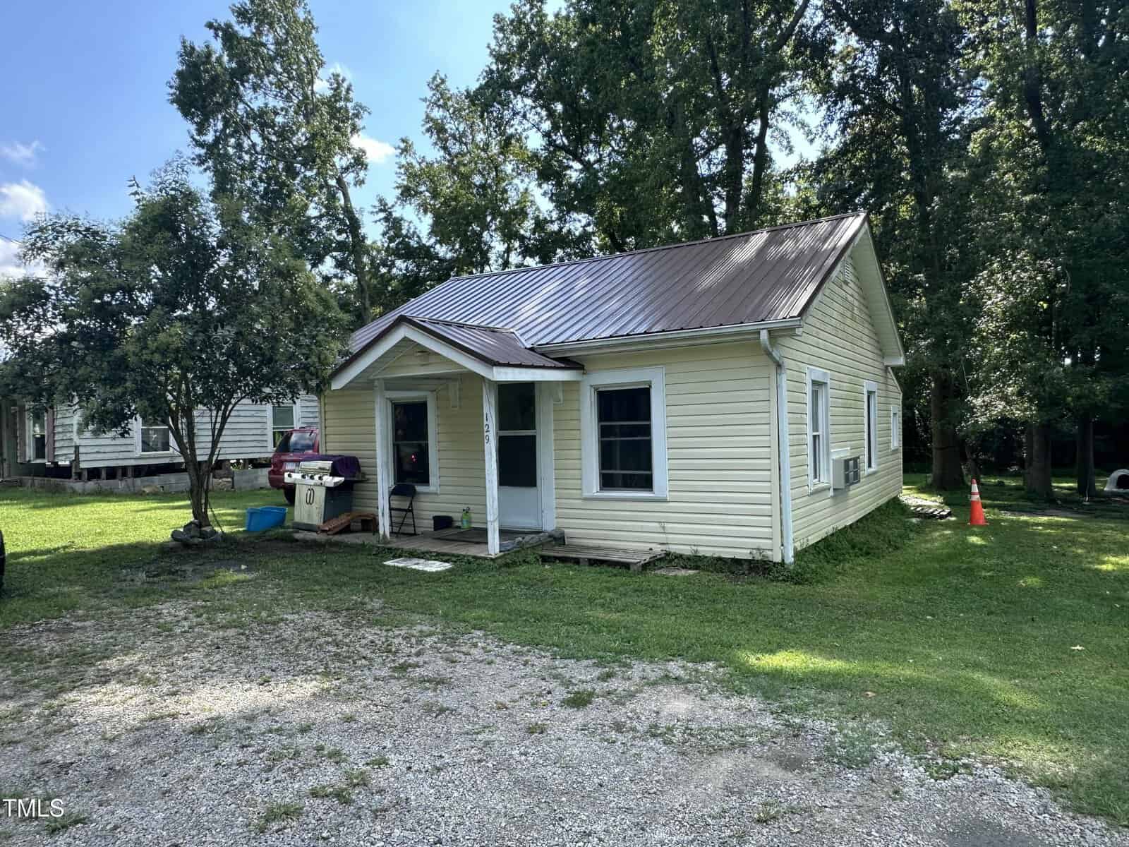 A small, single-story yellow house with white trim and a metal roof sits on a grassy lot at 129 Spring Valley Road, Henderson. A tree and grill are near the front porch, with tall trees providing shade in the background.