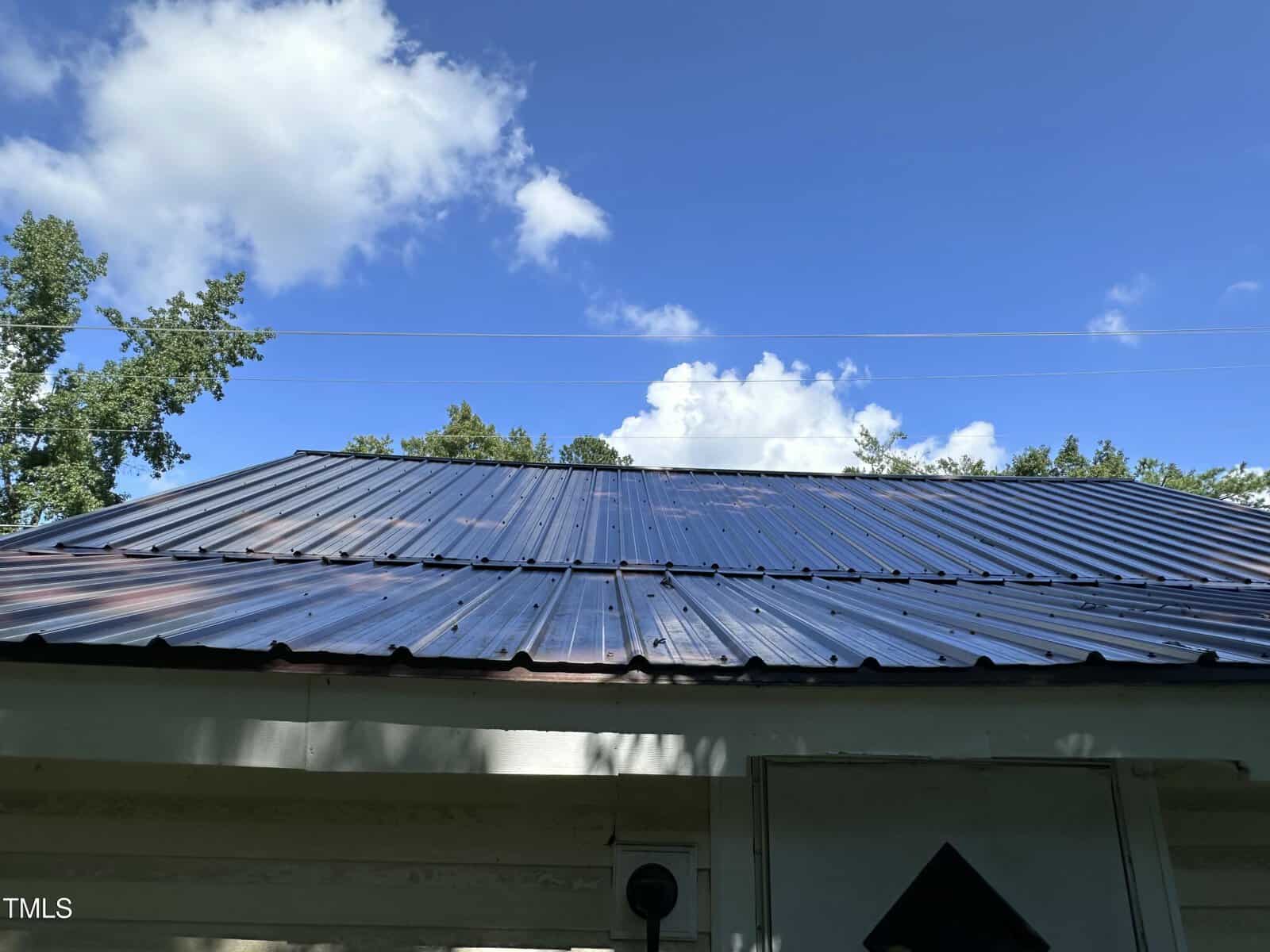 A close-up view of a metal roof on a building, photographed from below with a clear blue sky and a few clouds in the background. Trees and utility wires are visible behind the roof.