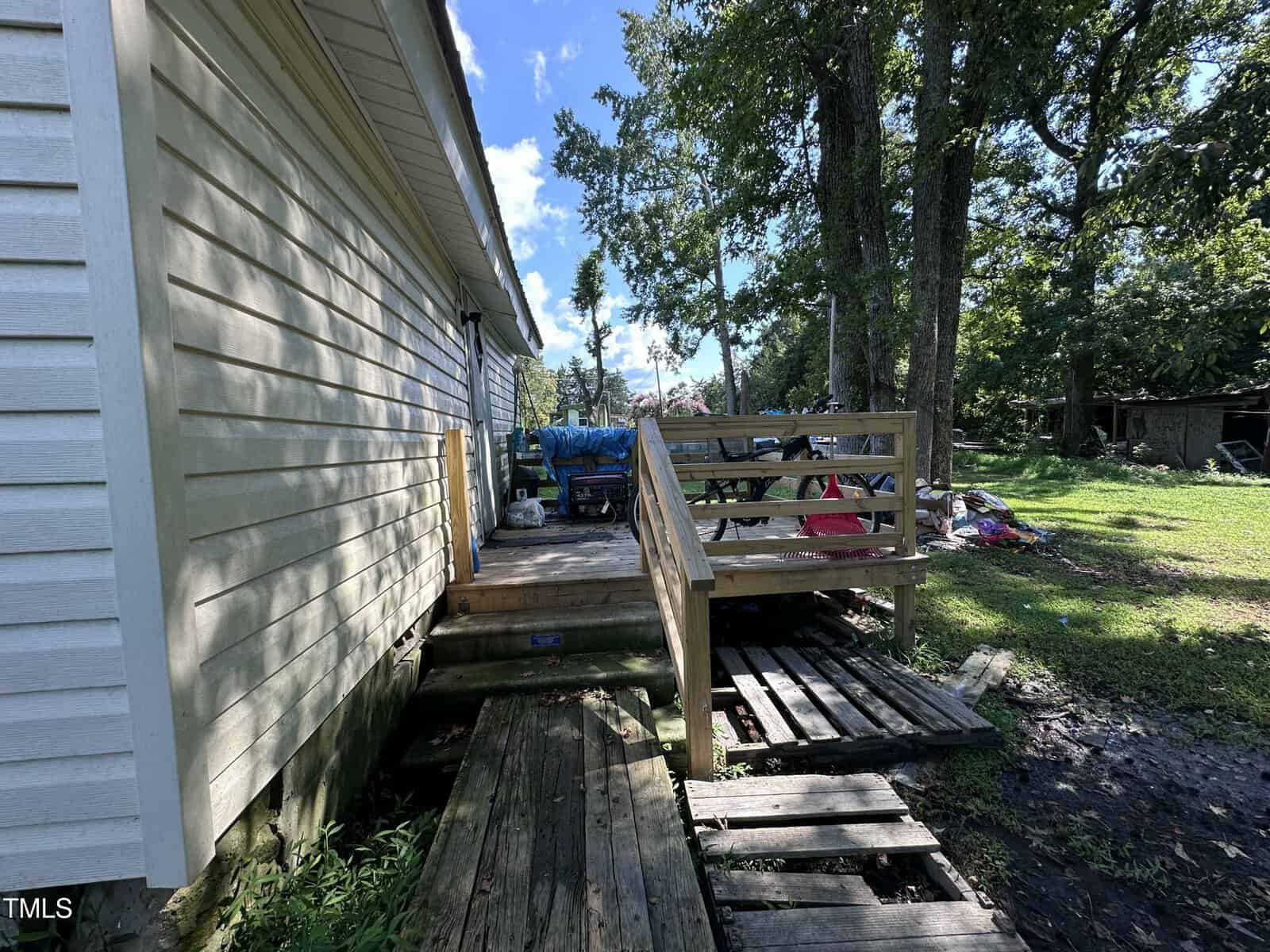 A wooden ramp and steps lead to a small deck attached to the side of a light-colored house. The yard is grassy with scattered items, and trees provide shade in the background under a blue sky.