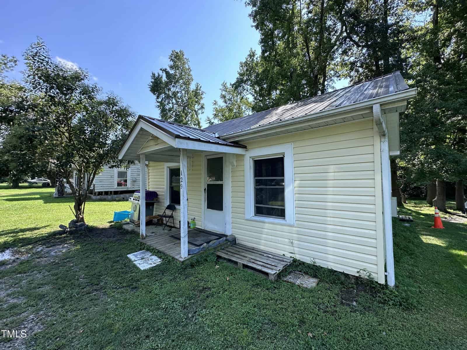 Small yellow house with a metal roof, white trim, and front porch on a grassy yard. Trees surround the home, and outdoor items sit by the porch. Another house is partially visible in the background.