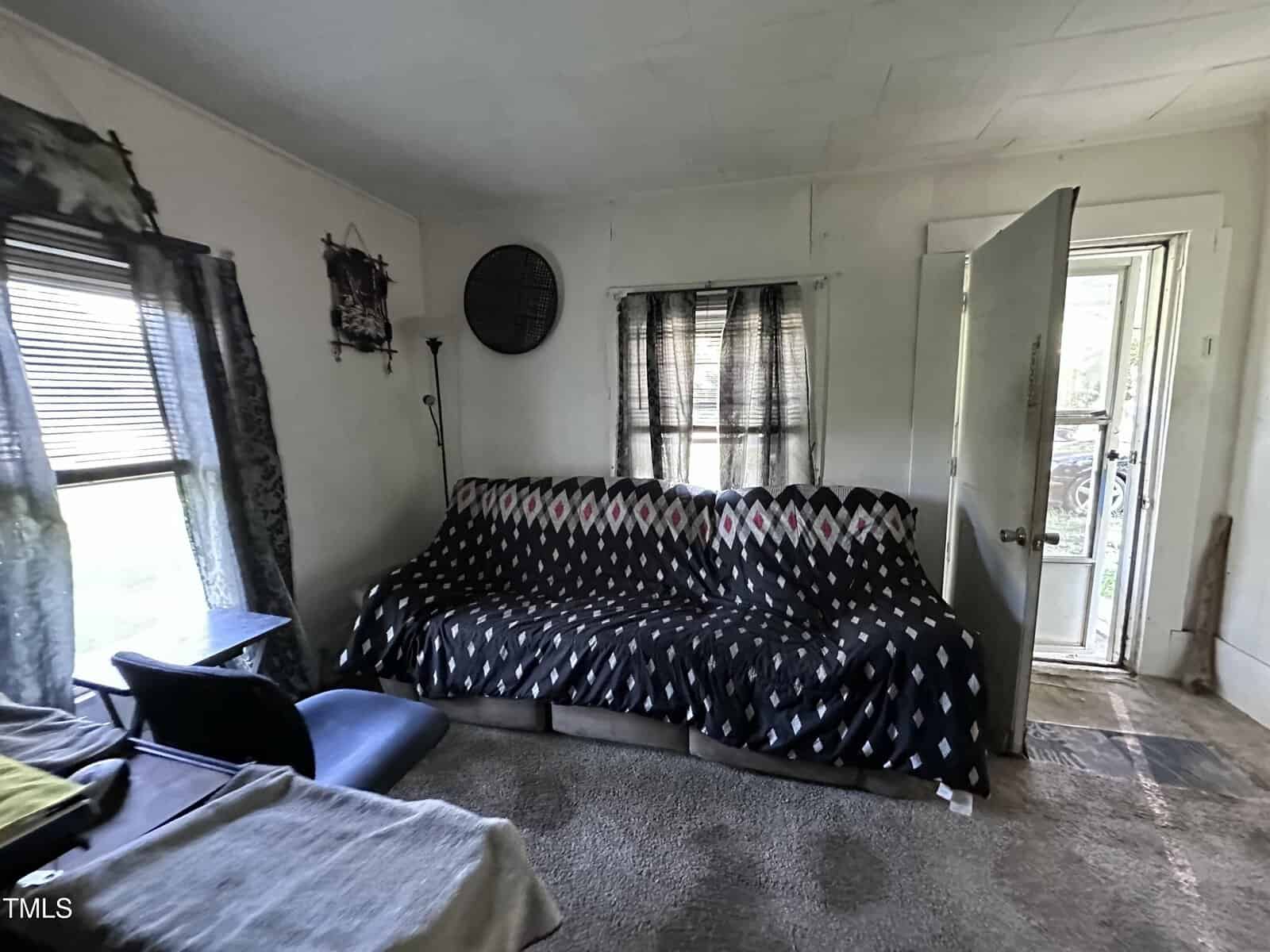 A small living room with a couch covered in a black patterned blanket, a window with curtains, an open door letting in light, and various furniture including a chair and small table. The carpet and walls show signs of wear.