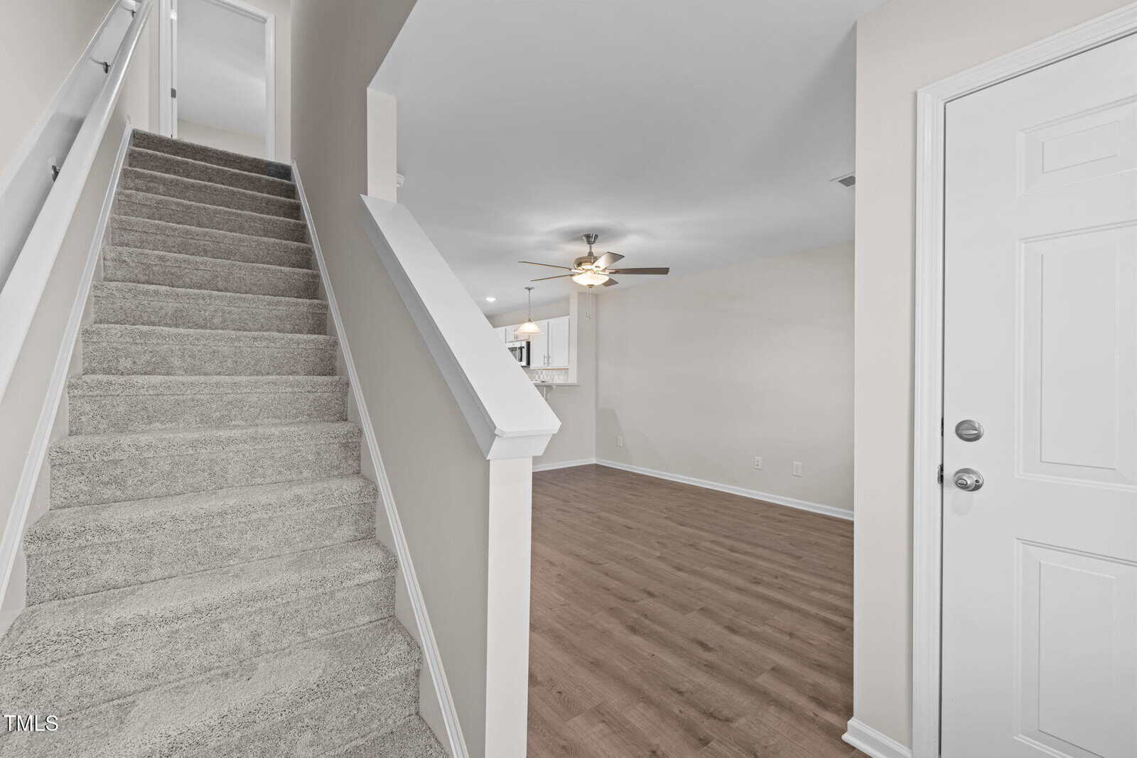 A view of a home interior showing carpeted stairs on the left, a white entry door on the right, and an open living area with wood flooring, neutral walls, and a ceiling fan in the background.