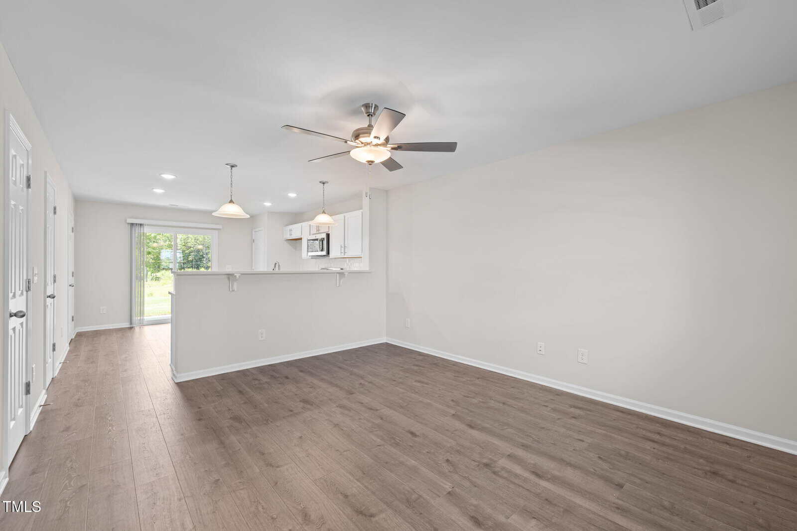 Unfurnished open-plan living and kitchen area with light-colored walls, wood-style flooring, ceiling fan, pendant lights above the kitchen counter, and sliding glass door leading outside.
