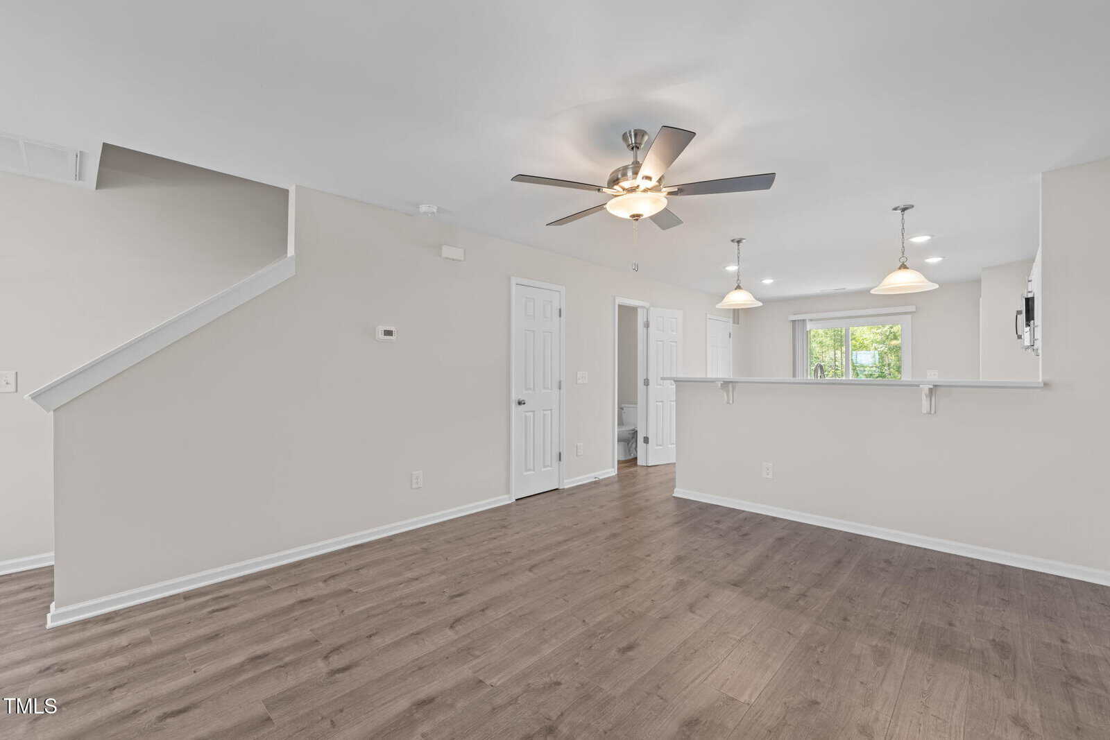Modern, unfurnished living area with wood floors, ceiling fan, and open view into a white kitchen with pendant lights. Stairs lead up on the left, and a glass door opens to the outside in the background.