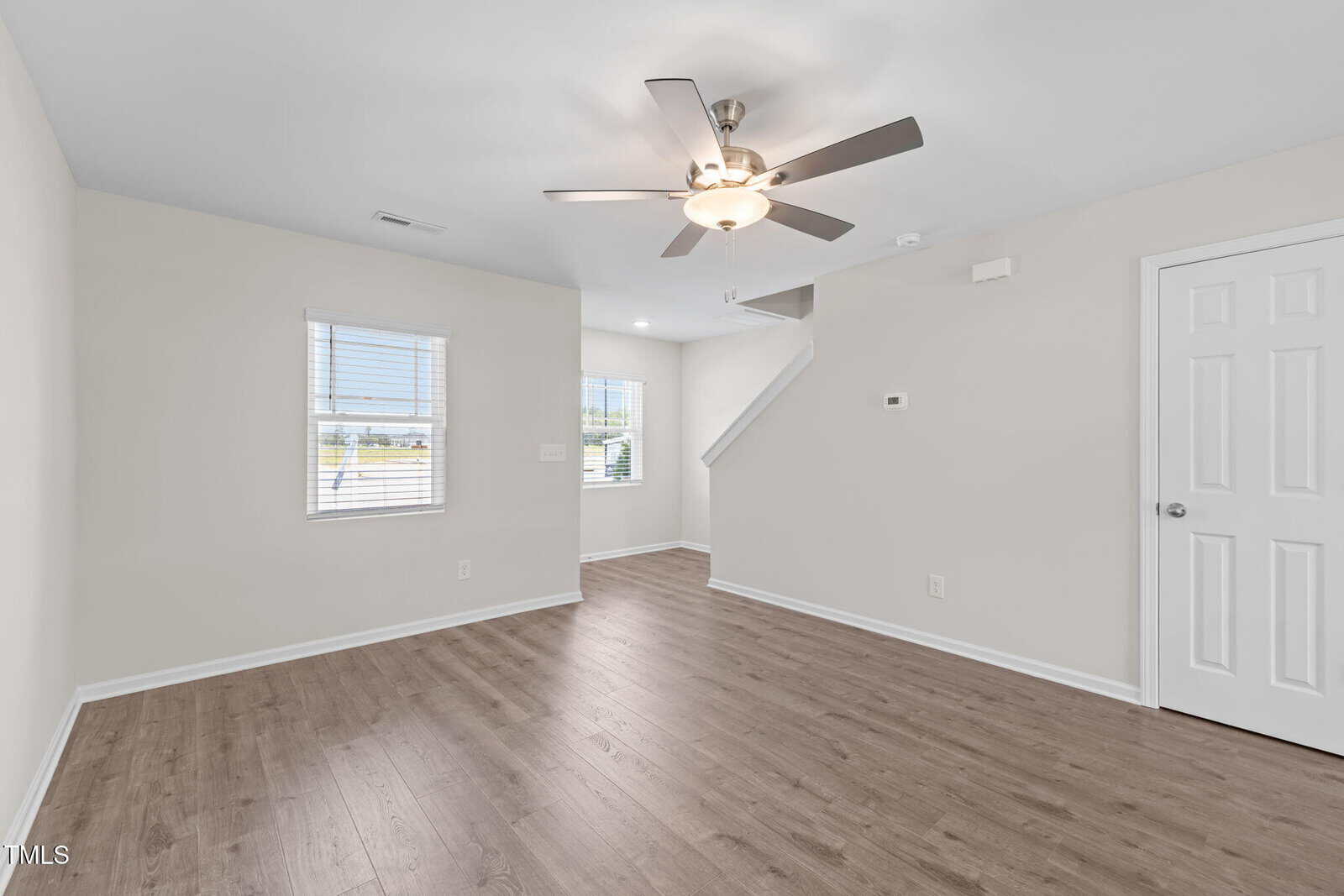 Bright, empty living room with light walls, wood flooring, ceiling fan with light, two windows, and a white door. Stairs with a white railing are visible in the background.