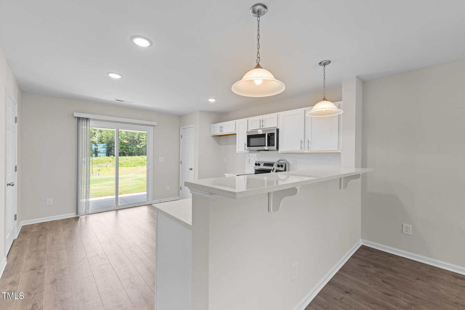 Open kitchen with white cabinets, stainless steel appliances, and pendant lighting. Bar counter faces a dining area with wood flooring. Glass sliding doors lead to a grassy backyard. Walls are light gray.