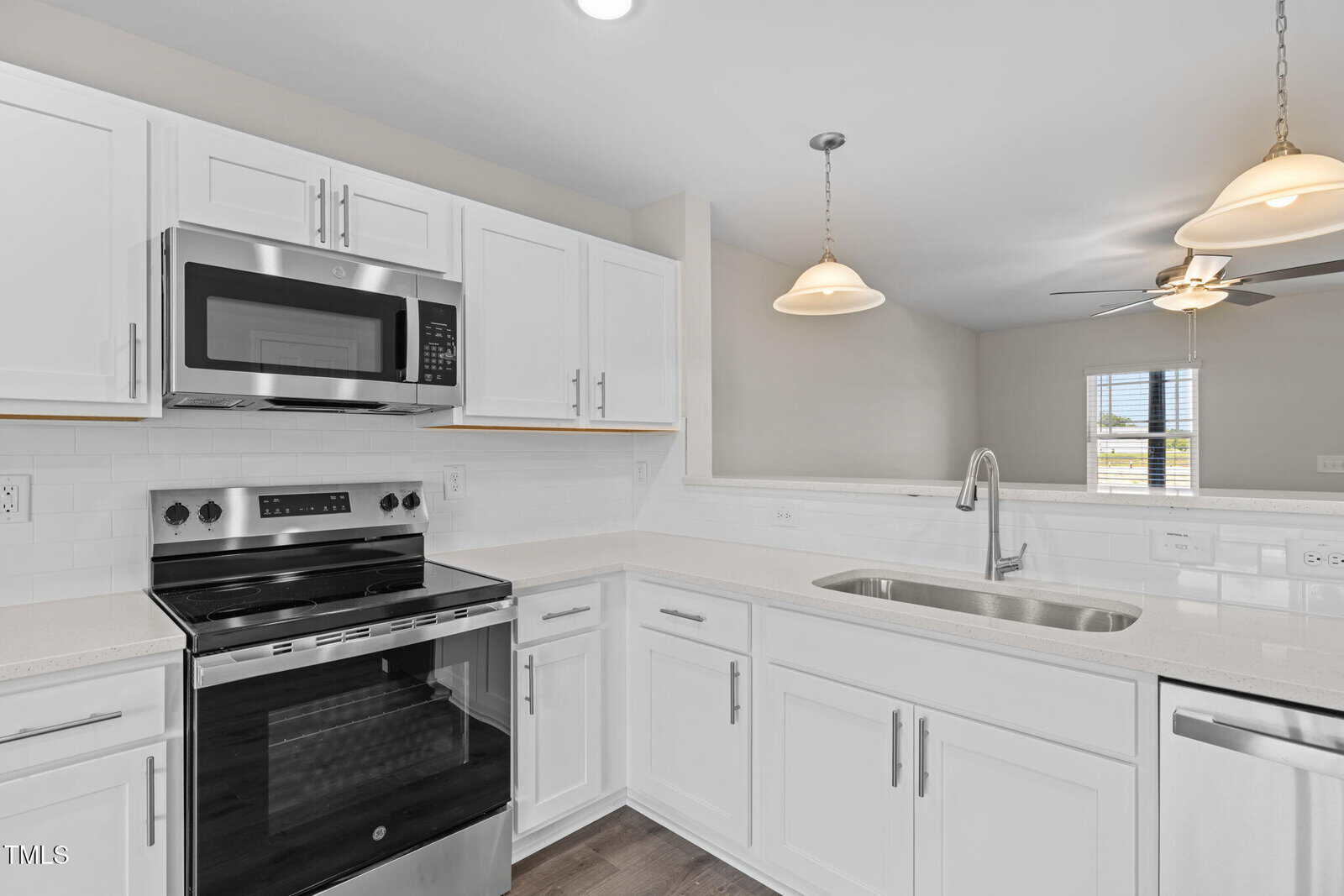 A modern kitchen with white cabinets, stainless steel appliances including an oven and microwave, a double sink, pendant lights, and a pass-through window to the living area with a ceiling fan.