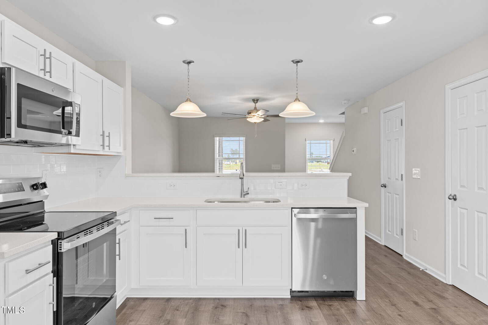 Modern kitchen with white cabinets, stainless steel appliances, a double sink under two pendant lights, and an open view into a living area with ceiling fan and windows. The floor is wood and walls are light-colored.