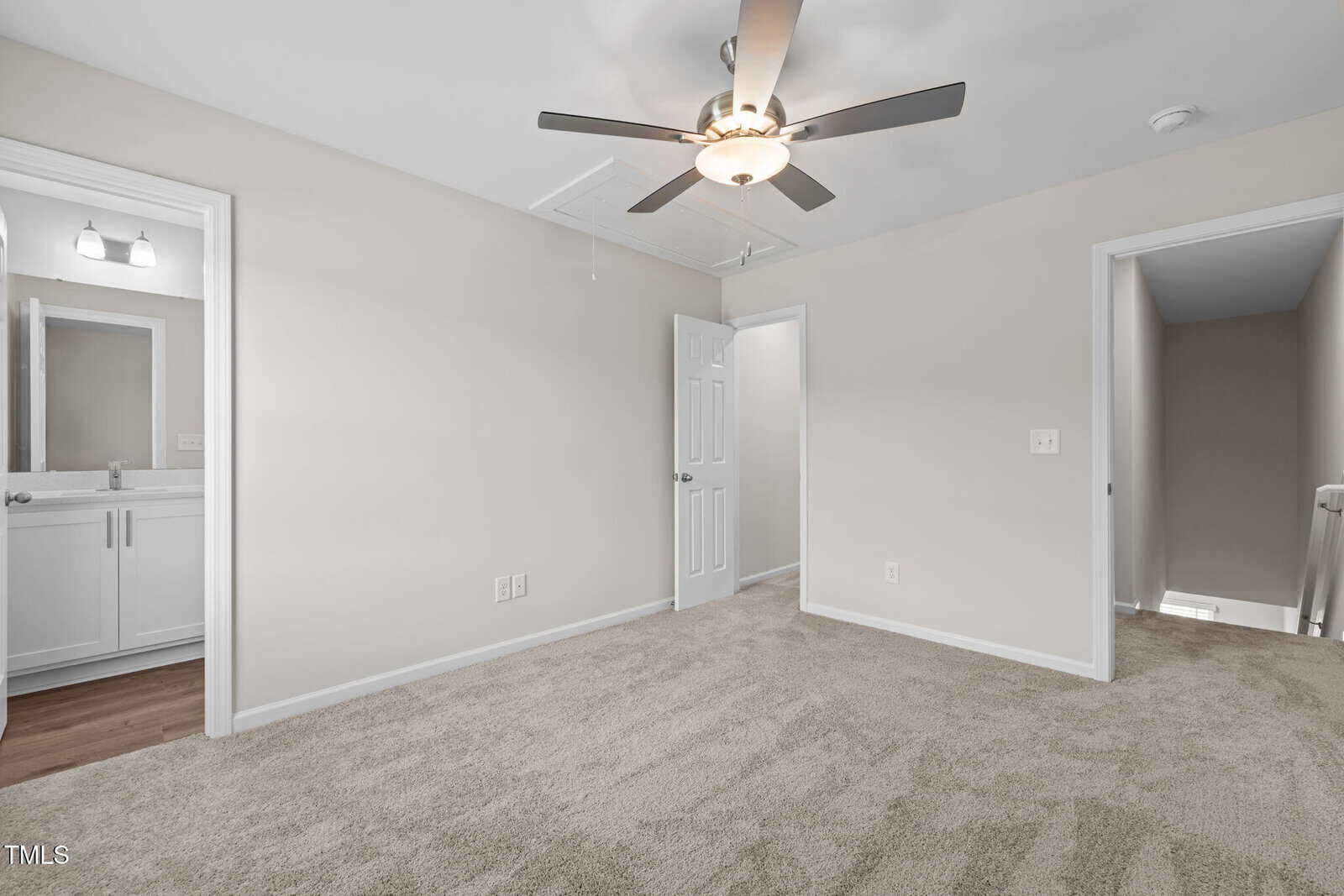 Empty carpeted bedroom with beige walls, ceiling fan with light, two doorways—one leading to a bathroom with a vanity, the other to a hallway and stairs. Natural light enters from the hallway.