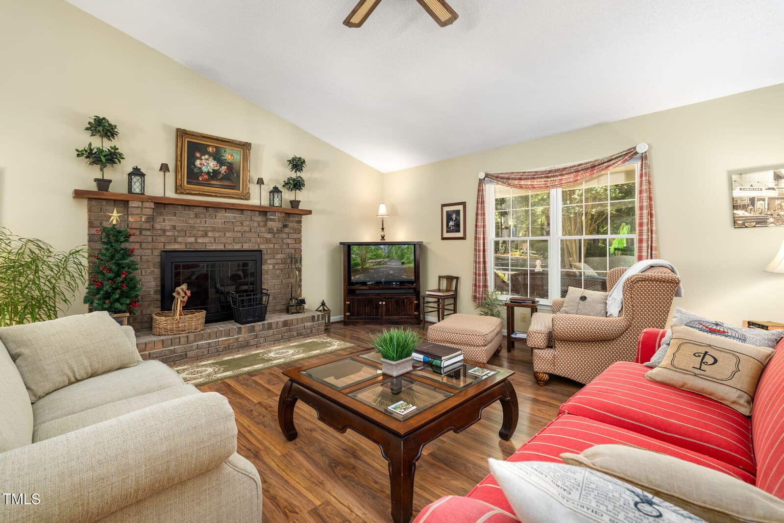 A living room with a brick fireplace, small decorated Christmas tree, beige and red sofas, glass coffee table, TV, armchair, wooden floors, large window with plaid curtains, and framed artwork on the walls.
