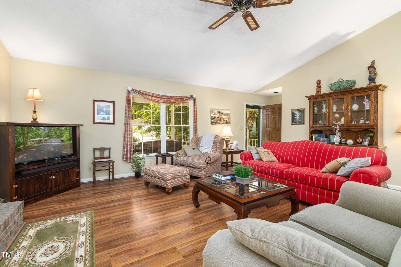 A living room with wood flooring, a red striped sofa, beige armchairs, a wooden coffee table, a wooden TV cabinet, a hutch with decorative items, and a large window with plaid curtains letting in natural light.