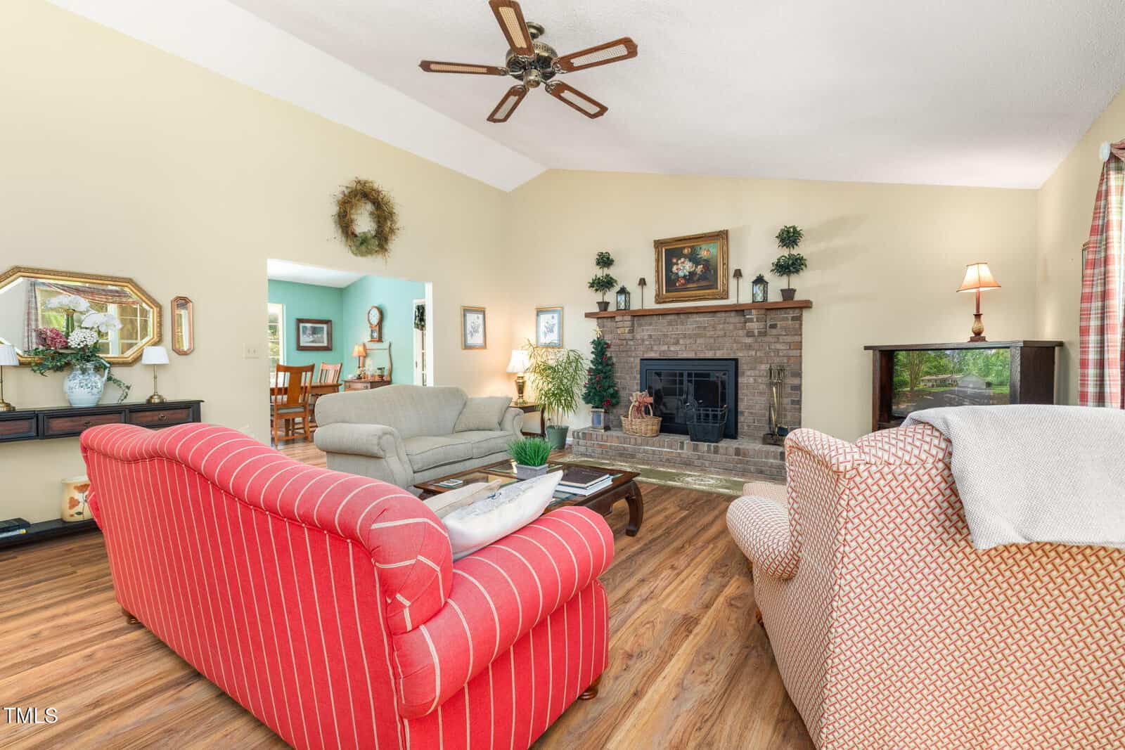 A living room with wood floors, a red striped couch, beige armchairs, a brick fireplace, and ceiling fan. Wall decor, plants, lamps, and a TV stand are also visible. An adjacent dining area can be seen through an open doorway.