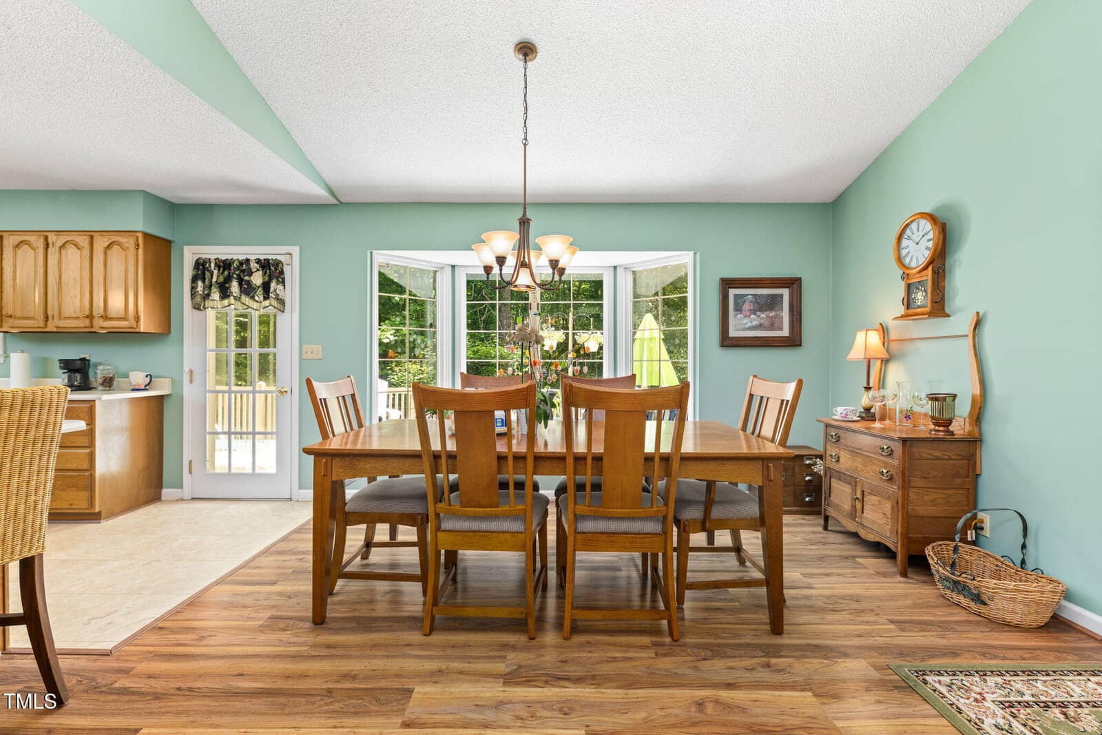 A dining room with a wooden table and six chairs, light blue-green walls, wood sideboard with a clock and lamp, and a chandelier. Large windows and a door lead to an outdoor area. The image also shows part of a kitchen.