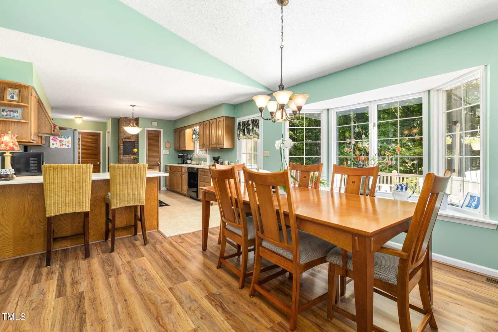 A spacious kitchen and dining area with wooden floors, a wooden dining table with eight chairs, a kitchen island with two wicker chairs, and large windows overlooking a green outdoor space.